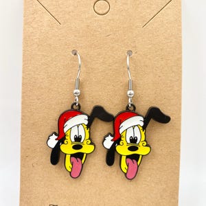 May include: A pair of dangle earrings featuring a cartoon dog with a yellow face, black ears, and a red Santa hat. The dog has a pink tongue sticking out. The earrings hang from silver hooks.
