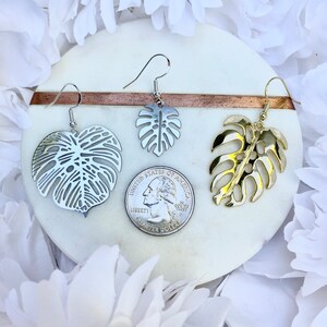 May include: Two pairs of earrings featuring a tropical leaf design. One pair is silver and the other is gold.