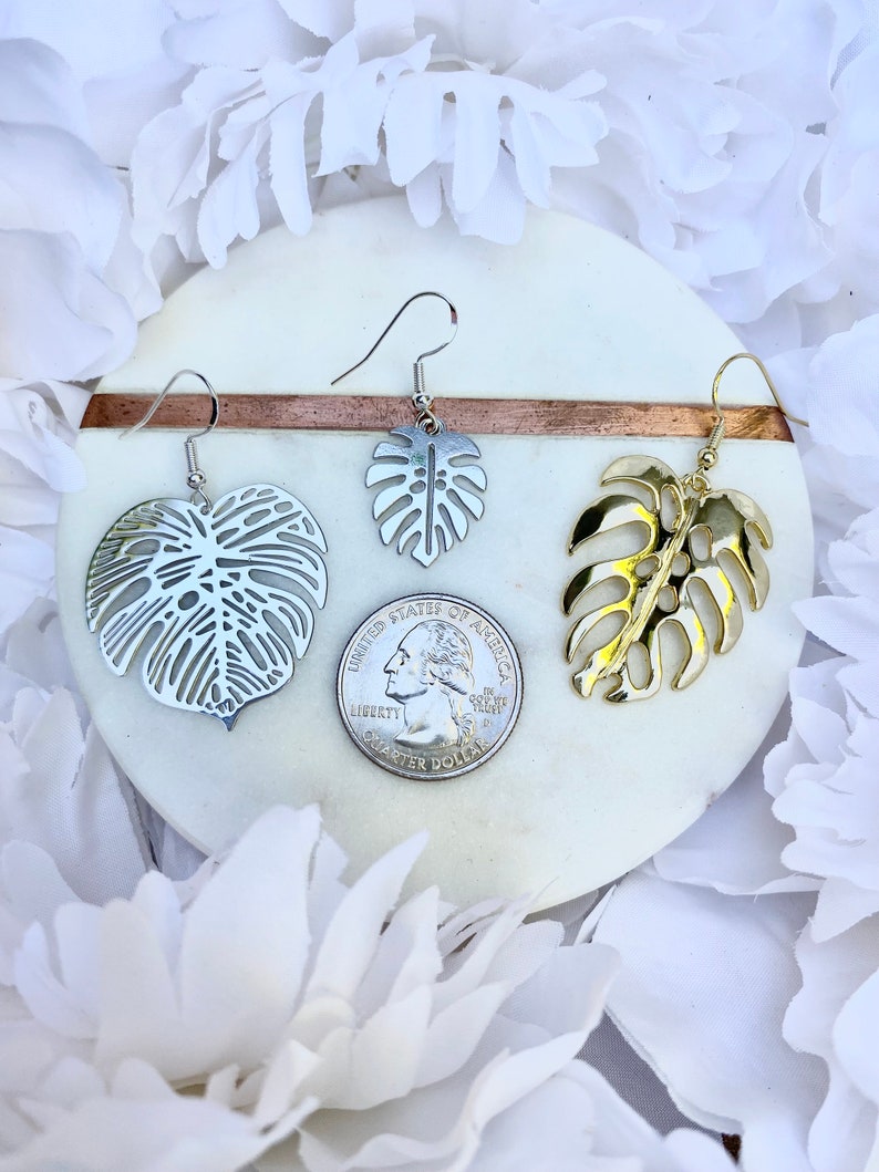 May include: Two pairs of earrings featuring a tropical leaf design. One pair is silver and the other is gold.