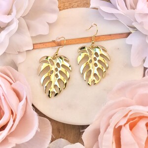 May include: A pair of gold-tone earrings featuring a cutout monstera leaf design. The earrings are displayed on a white surface with pink flowers.