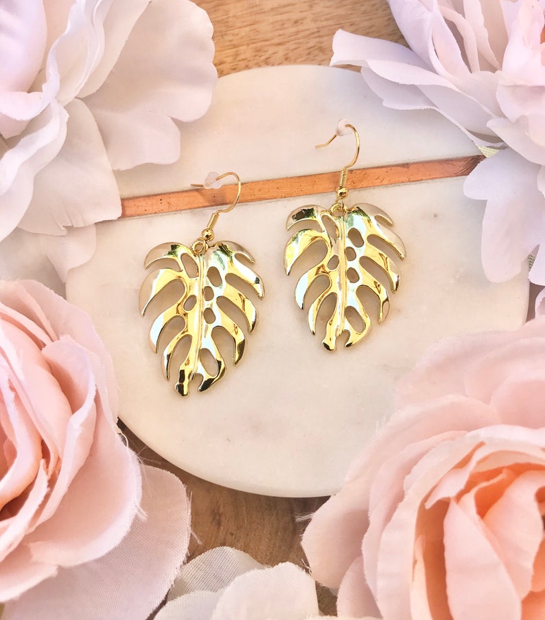 May include: A pair of gold-tone earrings featuring a cutout monstera leaf design. The earrings are displayed on a white surface with pink flowers.