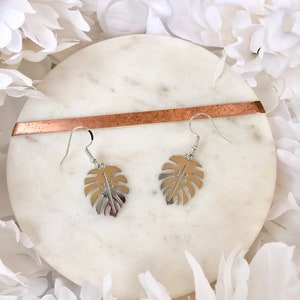 May include: A pair of silver earrings with a monstera leaf design. The earrings are on a white marble surface.
