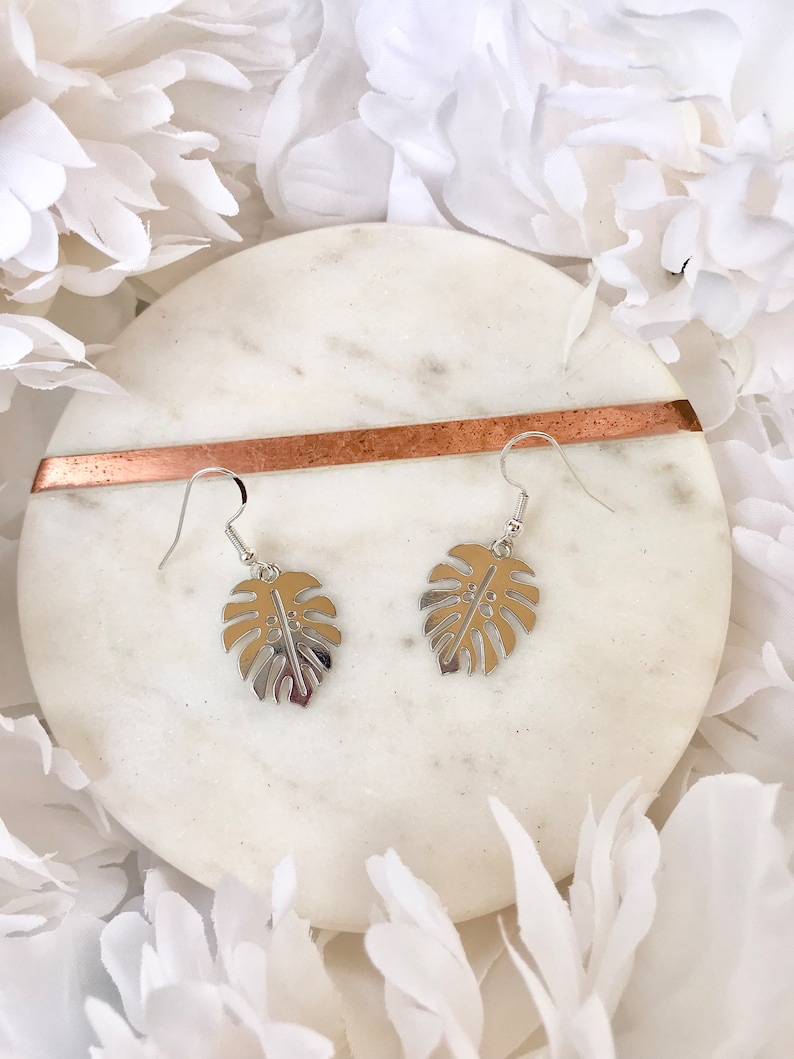 May include: A pair of silver earrings with a monstera leaf design. The earrings are on a white marble surface.