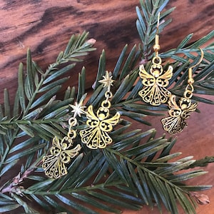 Puede incluir: Un par de pendientes de &aacute;ngel dorados con un dije de estrella en cada pendiente. Los pendientes est&aacute;n en una rama de pino verde.