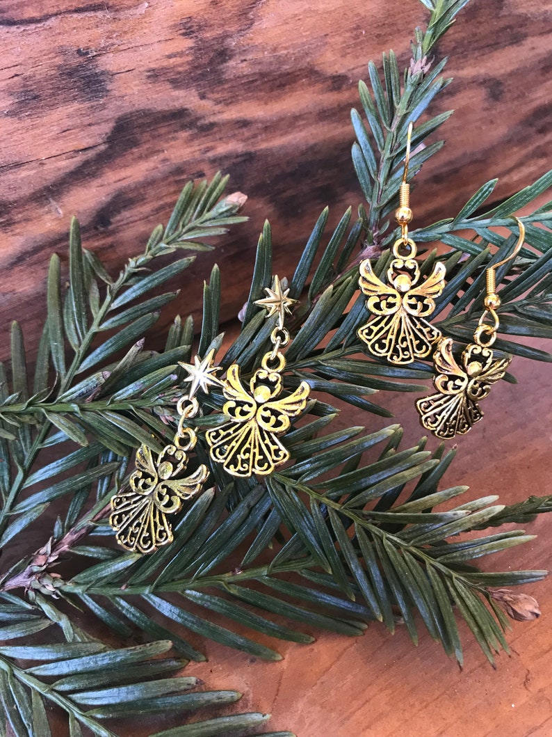 Puede incluir: Un par de pendientes de &aacute;ngel dorados con un dije de estrella en cada pendiente. Los pendientes est&aacute;n en una rama de pino verde.