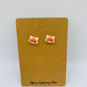 May include: A pair of small, white earrings featuring a cartoon red panda face with orange and white details. The earrings are displayed on a brown earring card with the text "Tiffany Epiphany Shop".