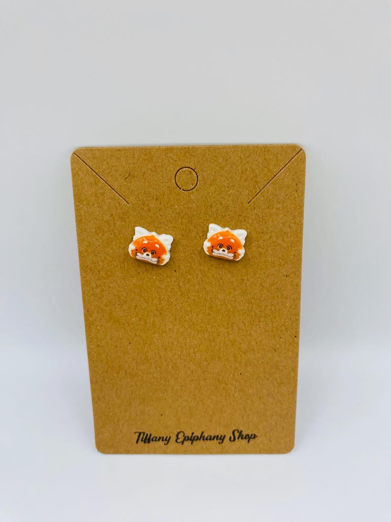 May include: A pair of small, white earrings featuring a cartoon red panda face with orange and white details. The earrings are displayed on a brown earring card with the text "Tiffany Epiphany Shop".