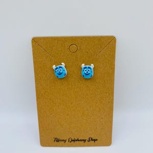 May include: A pair of blue monster-faced stud earrings on a brown card. The earrings have a light blue base with a smiling face and small horns. The card has the text "Tiffany Epiphany Shop" printed in black.