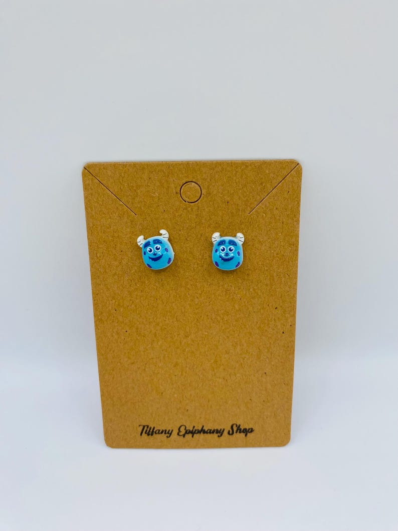 May include: A pair of blue monster-faced stud earrings on a brown card. The earrings have a light blue base with a smiling face and small horns. The card has the text "Tiffany Epiphany Shop" printed in black.
