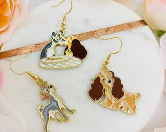 May include: A pair of gold-tone dangle earrings featuring the Disney characters Lady and the Tramp. One earring shows Lady and Tramp kissing, while the other earring shows Lady standing alone.