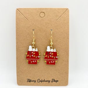 May include: A pair of gold-colored dangle earrings. Each earring features a red doghouse design with a white dog figure and a string of yellow and green lights. The earrings are displayed on a brown card with the text "Tiffany Epiphany Shop".