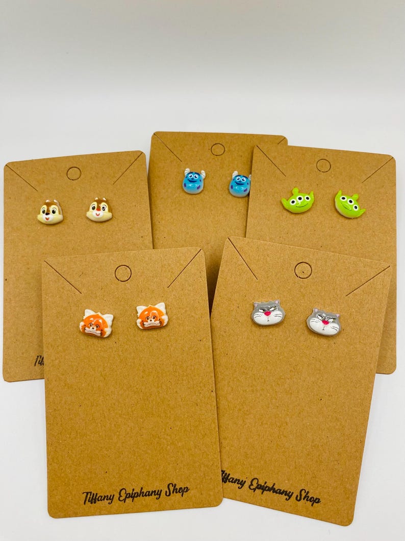 May include: Five pairs of novelty stud earrings on brown display cards. The earrings feature cartoon characters, including a chipmunk, blue aliens, green aliens, a red panda, and gray cats. The cards are labeled "Tiffany Epiphany Shop".