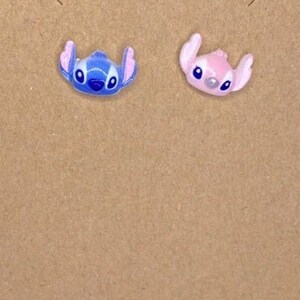 May include: A pair of stud earrings featuring the character Stitch from the Disney movie Lilo & Stitch. One earring is blue, the other is pink. The earrings are on a brown card with the text "Tiffany Epiphany Shop".