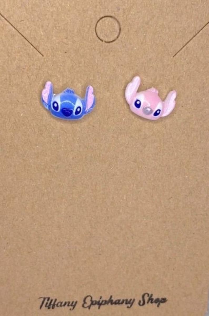 May include: A pair of stud earrings featuring the character Stitch from the Disney movie Lilo & Stitch. One earring is blue, the other is pink. The earrings are on a brown card with the text "Tiffany Epiphany Shop".