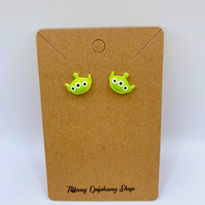 May include: A pair of lime green alien-shaped stud earrings with three white eyes each. The earrings are displayed on a brown cardboard earring card. The card has the text "Tiffany Epiphany Shop" printed in black.