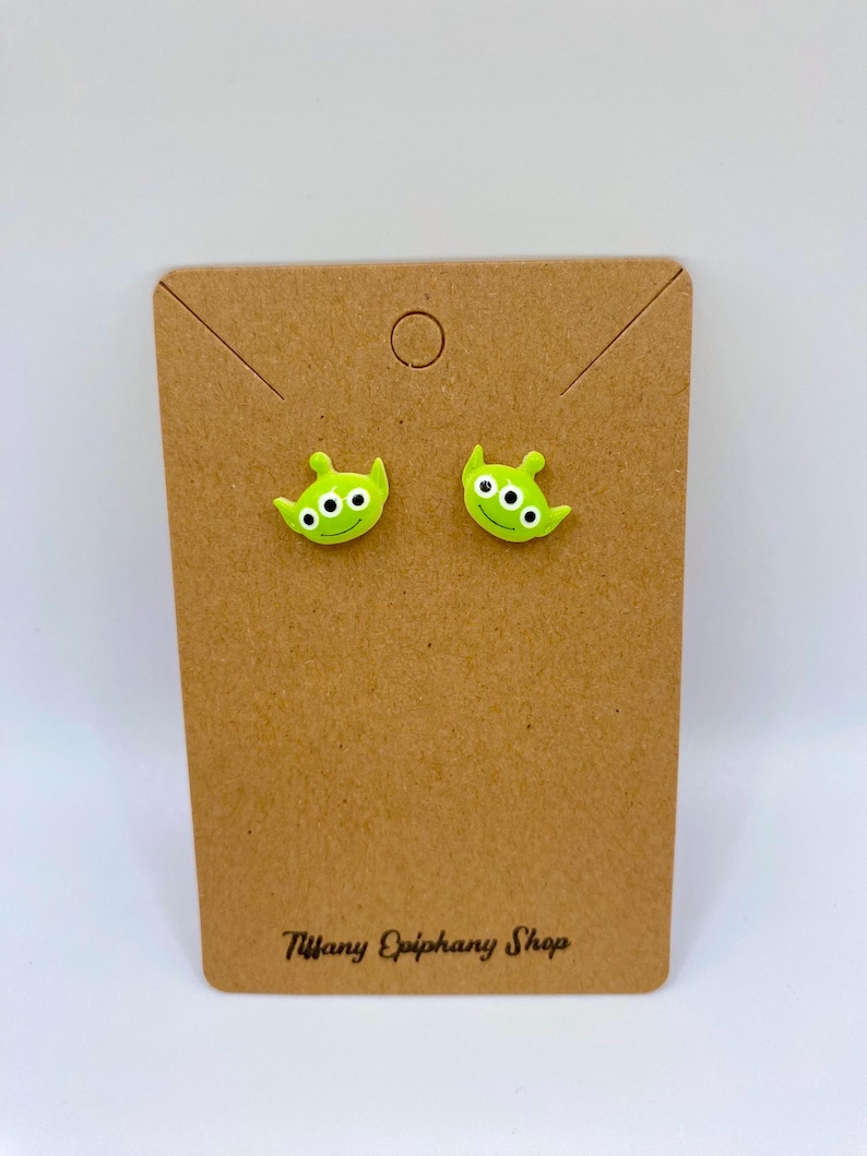 May include: A pair of lime green alien-shaped stud earrings with three white eyes each. The earrings are displayed on a brown cardboard earring card. The card has the text "Tiffany Epiphany Shop" printed in black.