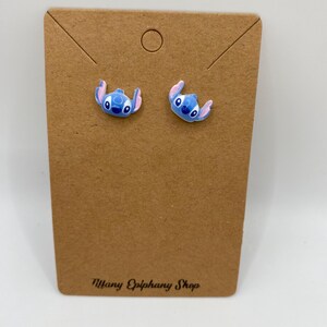 May include: A pair of blue and pink character stud earrings on a brown earring display card. The earrings feature a cartoon character design. The card has the text "Tiffany Epiphany Shop" printed on it.