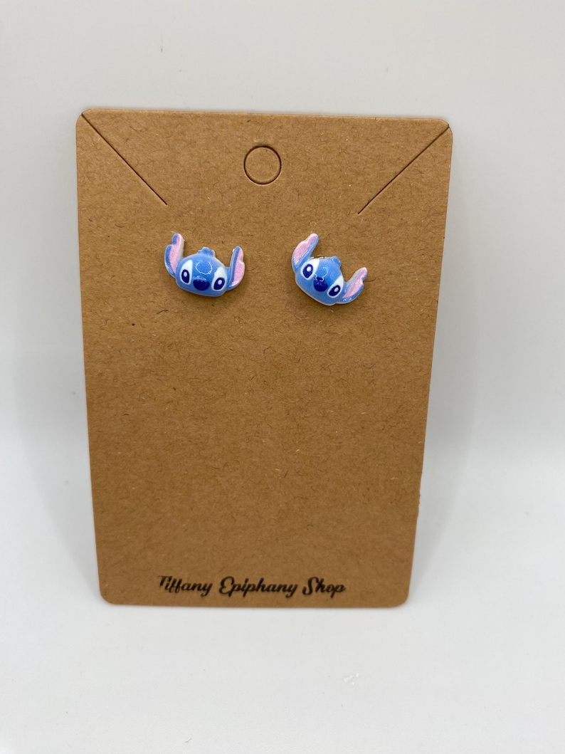 May include: A pair of blue and pink character stud earrings on a brown earring display card. The earrings feature a cartoon character design. The card has the text "Tiffany Epiphany Shop" printed on it.