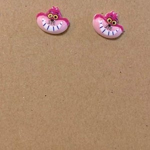 May include: A pair of pink Cheshire Cat earrings. Each earring features the cat's smiling face with a pink body, purple ears, and yellow eyes. The earrings are displayed on a brown card with the text "Tiffany Epiphany Shop".