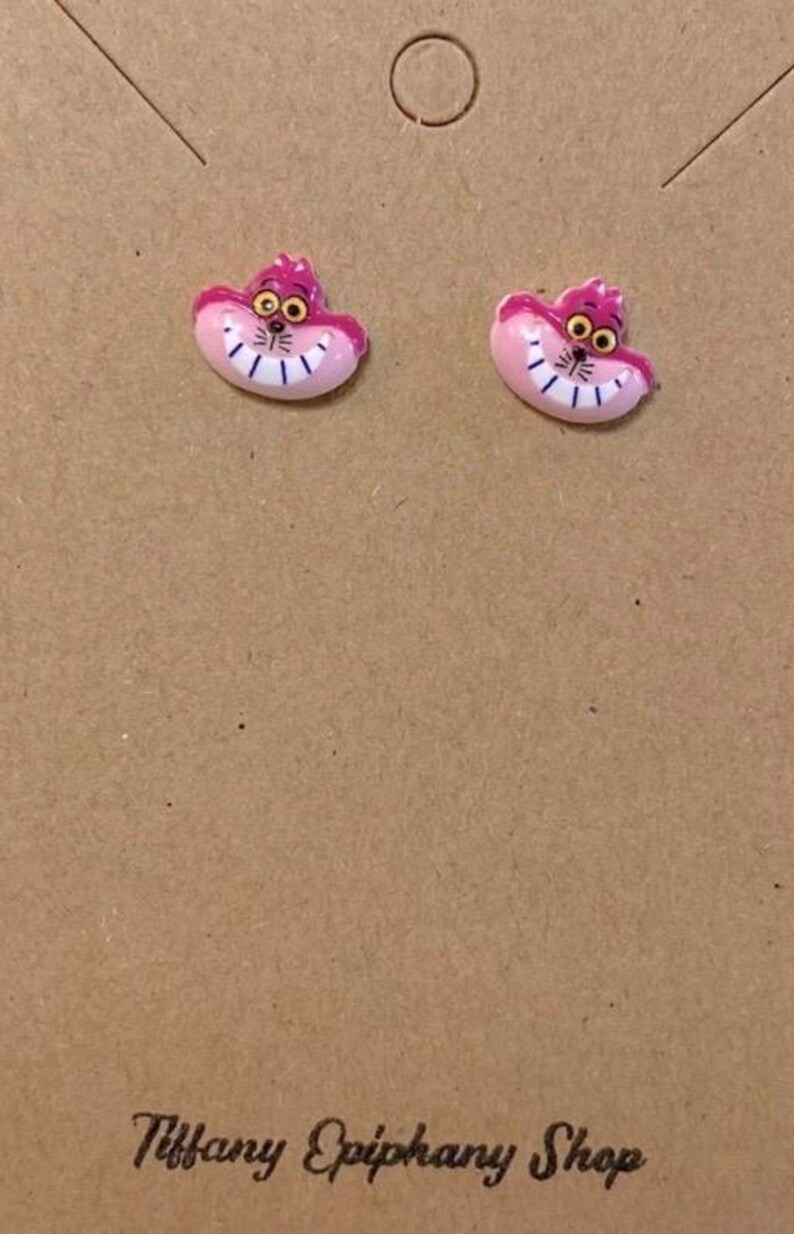 May include: A pair of pink Cheshire Cat earrings. Each earring features the cat's smiling face with a pink body, purple ears, and yellow eyes. The earrings are displayed on a brown card with the text "Tiffany Epiphany Shop".