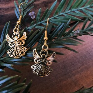 Puede incluir: Un par de pendientes de &aacute;ngel de oro con detalles intrincados. Los pendientes cuelgan de una rama de pino verde.