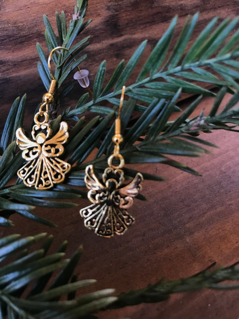 Puede incluir: Un par de pendientes de &aacute;ngel de oro con detalles intrincados. Los pendientes cuelgan de una rama de pino verde.