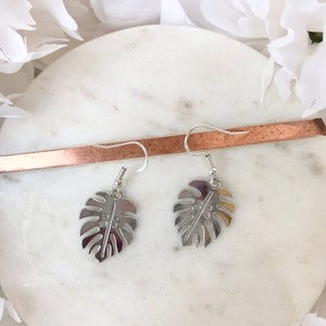 May include: A pair of silver earrings with a monstera leaf design. The earrings are hanging from a white marble surface.