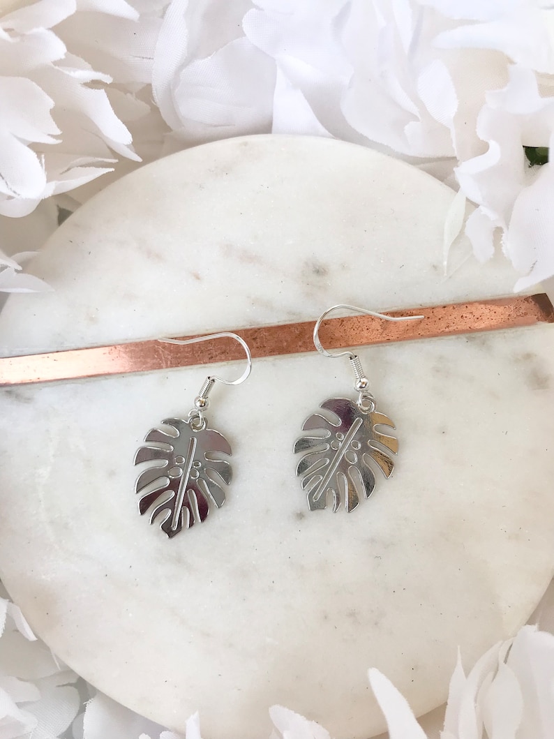 May include: A pair of silver earrings with a monstera leaf design. The earrings are hanging from a white marble surface.