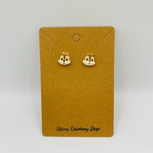 May include: A pair of stud earrings featuring Chip from Chip 'n Dale Rescue Rangers. The earrings are light brown with white and red accents, set on a brown card with the text "Tiffany Epiphany Shop".