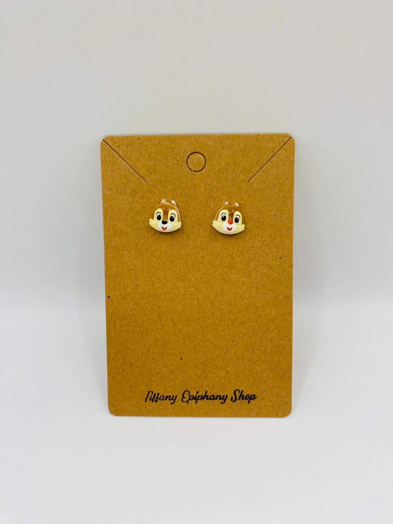 May include: A pair of stud earrings featuring Chip from Chip 'n Dale Rescue Rangers. The earrings are light brown with white and red accents, set on a brown card with the text "Tiffany Epiphany Shop".