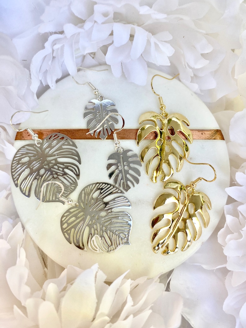 May include: Two pairs of earrings featuring a tropical leaf design. One pair is silver and the other is gold.