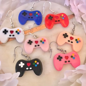 Puede incluir: Seis pares de pendientes de mando de videojuegos de colores. Los pendientes están hechos de plástico y tienen un diseño de corazón en la parte delantera. Los mandos son de color azul, rojo, blanco, rosa, negro y rosa claro.