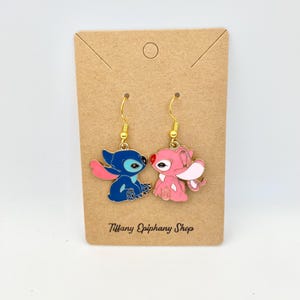 Op de afbeelding: Een paar emaille oorbellen met blauwe en roze cartoonfiguren met goudkleurige haken. De oorbellen worden gepresenteerd op een bruine kaart met de tekst "Tiffany Epiphany Shop".
