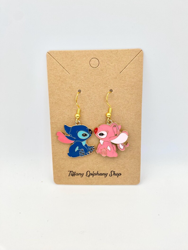 Op de afbeelding: Een paar emaille oorbellen met blauwe en roze cartoonfiguren met goudkleurige haken. De oorbellen worden gepresenteerd op een bruine kaart met de tekst "Tiffany Epiphany Shop".