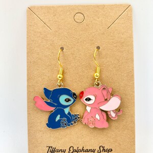 Op de afbeelding: Een paar goudkleurige oorhangers met cartoonfiguren. E&eacute;n oorbel is blauw met roze accenten, de andere is roze. De oorbellen zitten op een bruine kaart met de tekst "Tiffany Epiphany Shop".