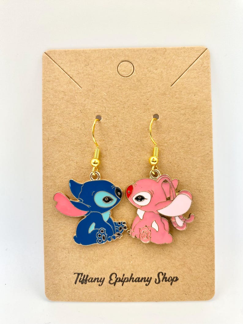 Op de afbeelding: Een paar goudkleurige oorhangers met cartoonfiguren. E&eacute;n oorbel is blauw met roze accenten, de andere is roze. De oorbellen zitten op een bruine kaart met de tekst "Tiffany Epiphany Shop".