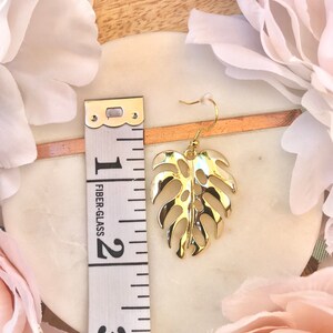 May include: Gold-tone monstera leaf dangle earrings with a hook closure. The earrings are approximately 2 inches long.
