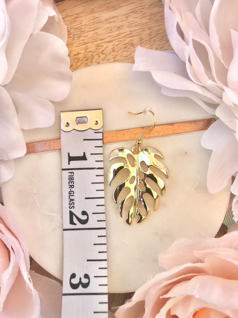 May include: Gold-tone monstera leaf dangle earrings with a hook closure. The earrings are approximately 2 inches long.