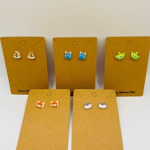 May include: A collection of stud earrings featuring cartoon character designs. The earrings are displayed on brown earring cards. The cards are labeled "Tiffany Epiphany Shop". The earrings include Chip and Dale, Mike Wazowski, the Little Green Men, a fox, and a cat.
