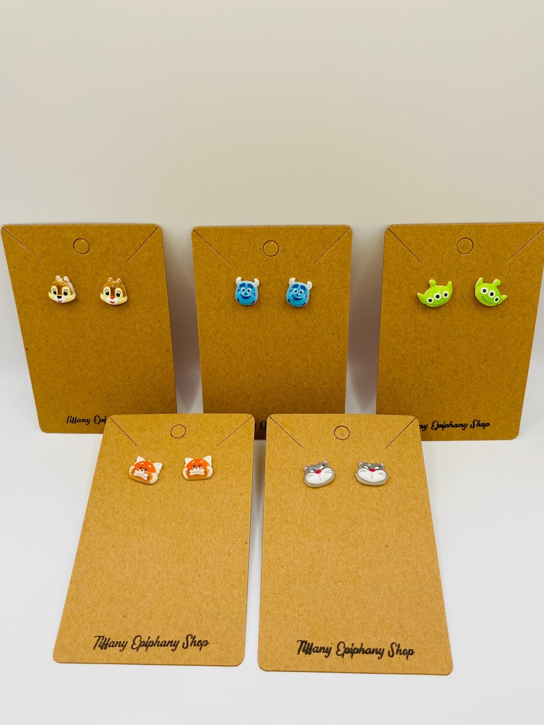May include: A collection of stud earrings featuring cartoon character designs. The earrings are displayed on brown earring cards. The cards are labeled "Tiffany Epiphany Shop". The earrings include Chip and Dale, Mike Wazowski, the Little Green Men, a fox, and a cat.