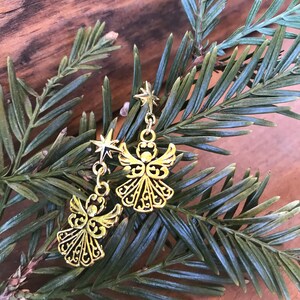 Puede incluir: Un par de pendientes de &aacute;ngel dorados con detalles de estrellas colgando de una rama de pino verde.