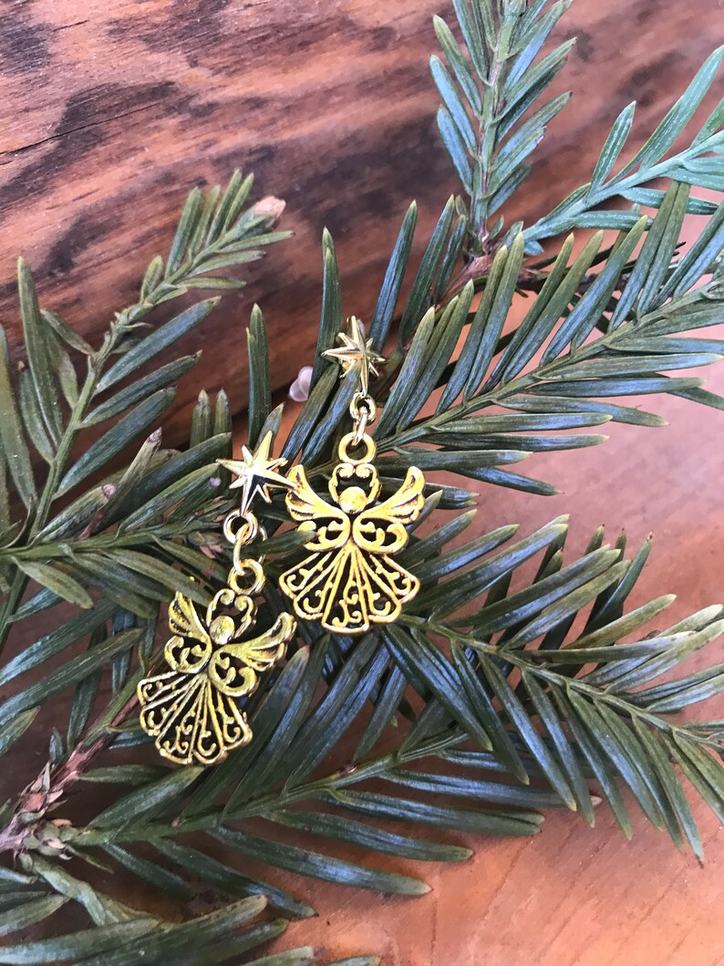 Puede incluir: Un par de pendientes de &aacute;ngel dorados con detalles de estrellas colgando de una rama de pino verde.