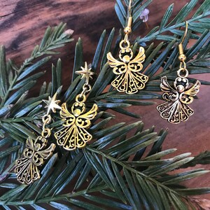 Puede incluir: Un par de pendientes de &aacute;ngel dorados con un dije de estrella en cada pendiente. Los pendientes est&aacute;n sobre un fondo de rama de pino verde.