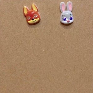 May include: A pair of stud earrings featuring cartoon fox and bunny faces. The fox has red and yellow details, while the bunny is gray with pink ears. The earrings are on a brown card with the text "Tiffany Epiphany Shop".