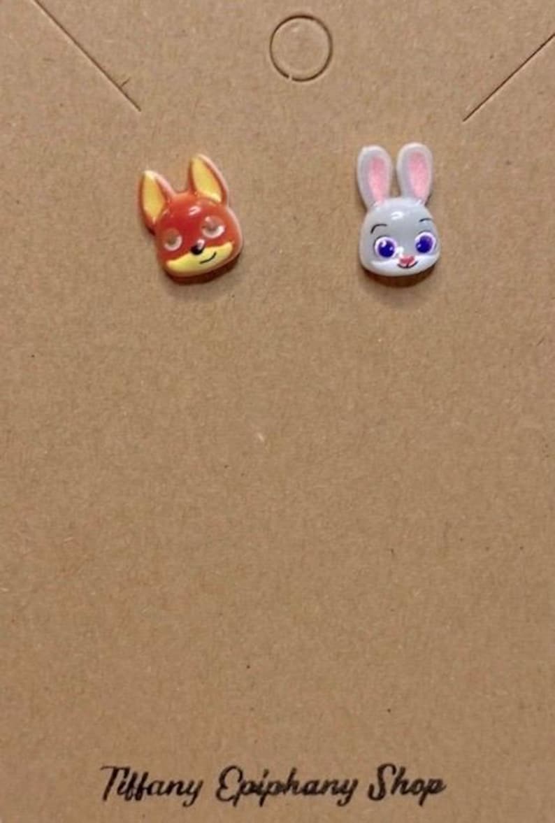 May include: A pair of stud earrings featuring cartoon fox and bunny faces. The fox has red and yellow details, while the bunny is gray with pink ears. The earrings are on a brown card with the text "Tiffany Epiphany Shop".