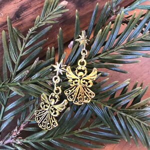 Puede incluir: Un par de pendientes de &aacute;ngel dorados con un dije de estrella colgando de cada uno. Los pendientes est&aacute;n en una rama de pino verde.