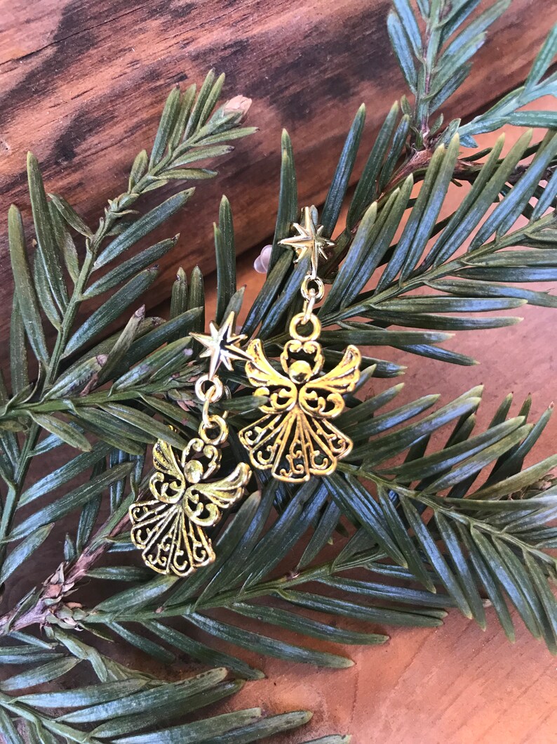 Puede incluir: Un par de pendientes de &aacute;ngel dorados con un dije de estrella colgando de cada uno. Los pendientes est&aacute;n en una rama de pino verde.