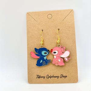 Op de afbeelding: Een paar goudkleurige oorhangers met blauwe en roze cartoonfiguren. Het blauwe personage heeft een roze buik en oren, terwijl het roze personage witte oren heeft. De oorbellen worden gepresenteerd op een bruine kaart met de tekst "Tiffany Epiphany Shop".