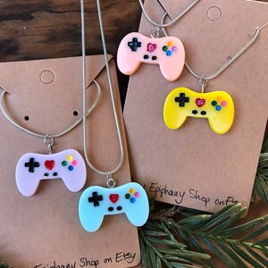 Peut inclure: Quatre colliers en argent avec des pendentifs de manette de jeu en plastique coloré. Les manettes sont rose, jaune, bleu clair et violet. Chaque manette a un signe plus noir, un cœur rouge et des cercles colorés. Les colliers sont présentés sur du carton brun avec le texte "Tiny Epiphany Shop on Etsy".
