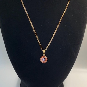 May include: A gold-colored necklace with a pendant featuring a red, white, and blue shield design. The pendant is round with a star in the center. The necklace is displayed on a black mannequin.